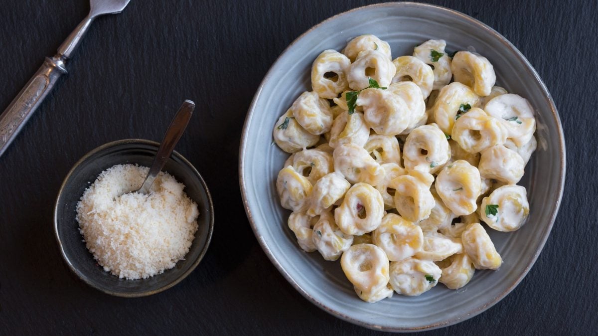Tortellini in Knoblauch-Sahne-Sauce auf hellem Teller, angerichtet auf dunklem Untergrund. Daneben steht eine Schale mit geriebenem Käse.