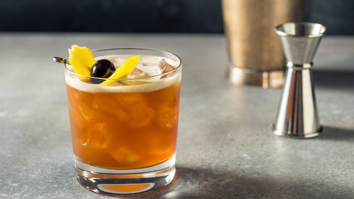 Amaretto Sour mit Zitronenzeste und Cocktailkirsche im Old Fashioned Glas auf grauem Tisch. Im Hintergrund Barmaß und Shaker. Frontalansicht.