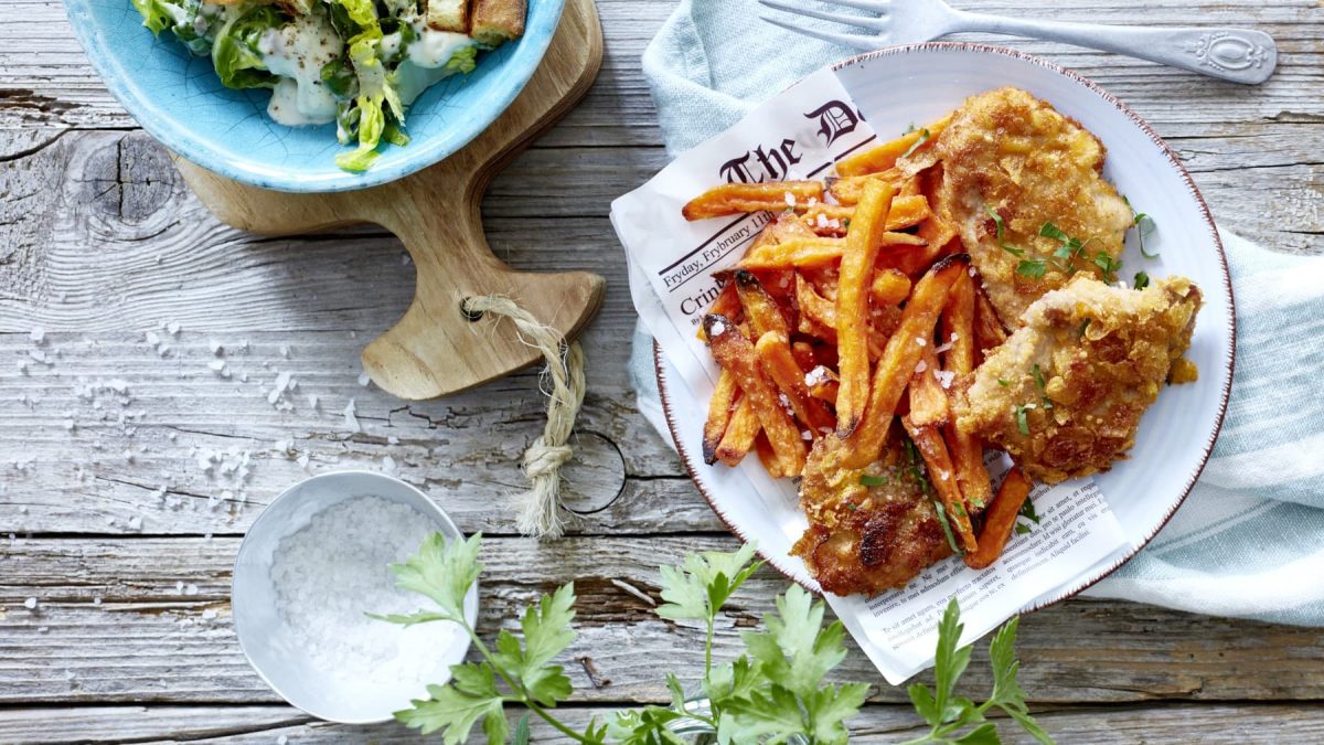 Amerikanische Crisp-Schnitzel mit Süßkartoffel-Pommes auf einem Teller neben einer Schale Salz und einem Teller mit Salat vor Holzhintergrund