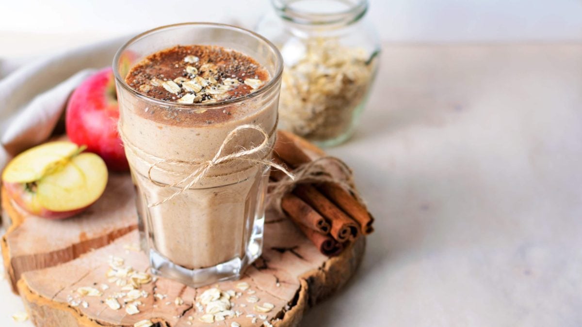 Ein Glas mit dem Apfel-Zimt-Smoothie auf einer Holzscheibe mit frischen Äpfeln und Haferflocken im Hintergrund.