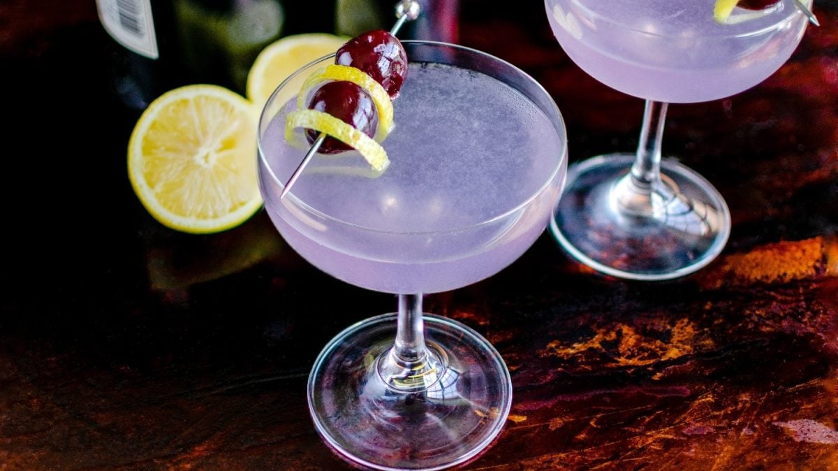 Ein Coupette-Glas mit dem violetten Aviation auf einem dunklem Holzuntergrund dekoriert mit Kirschen und einer Zitronenschale.