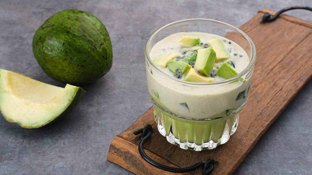 Avocado und Kondensmilch in einem Glas mit Basilikumsamen auf einem Holzbrett und grauem Untergrund. Im Hintergrund eine angeschnittene Avocado.