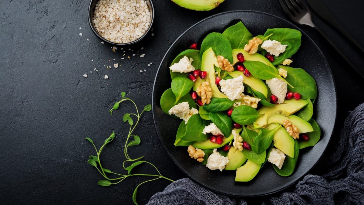 Ein schwarzer Teller mit Avocado-Salat, auf schwarzem Hintergrund neben Besteck und frischen Zutaten.