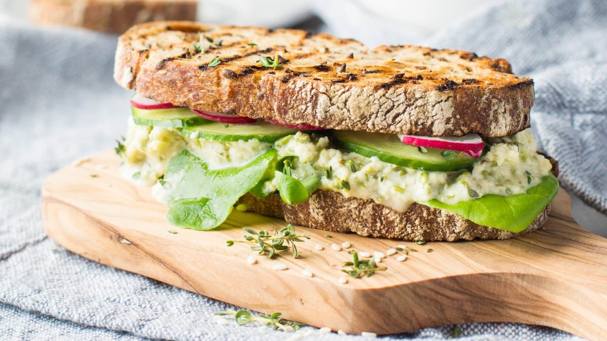 Ein belegtes Sandwich mit Bohnen-Hummus auf einem hellen Holzbrett und einer hellblauen Decke als Hintergrund.