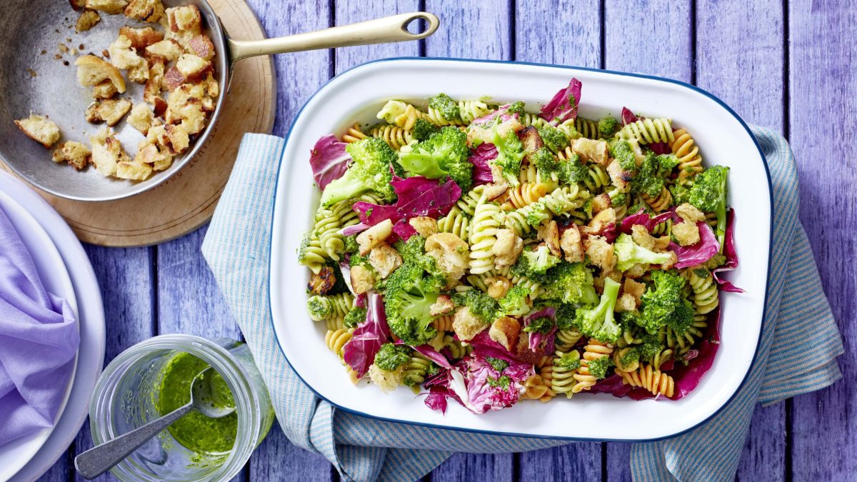 Schüssel mit Broccoli-Nudelsalat neben Schüssel mit Brotbröseln und Glas mit Pesto auf blauen Holzhintergrund
