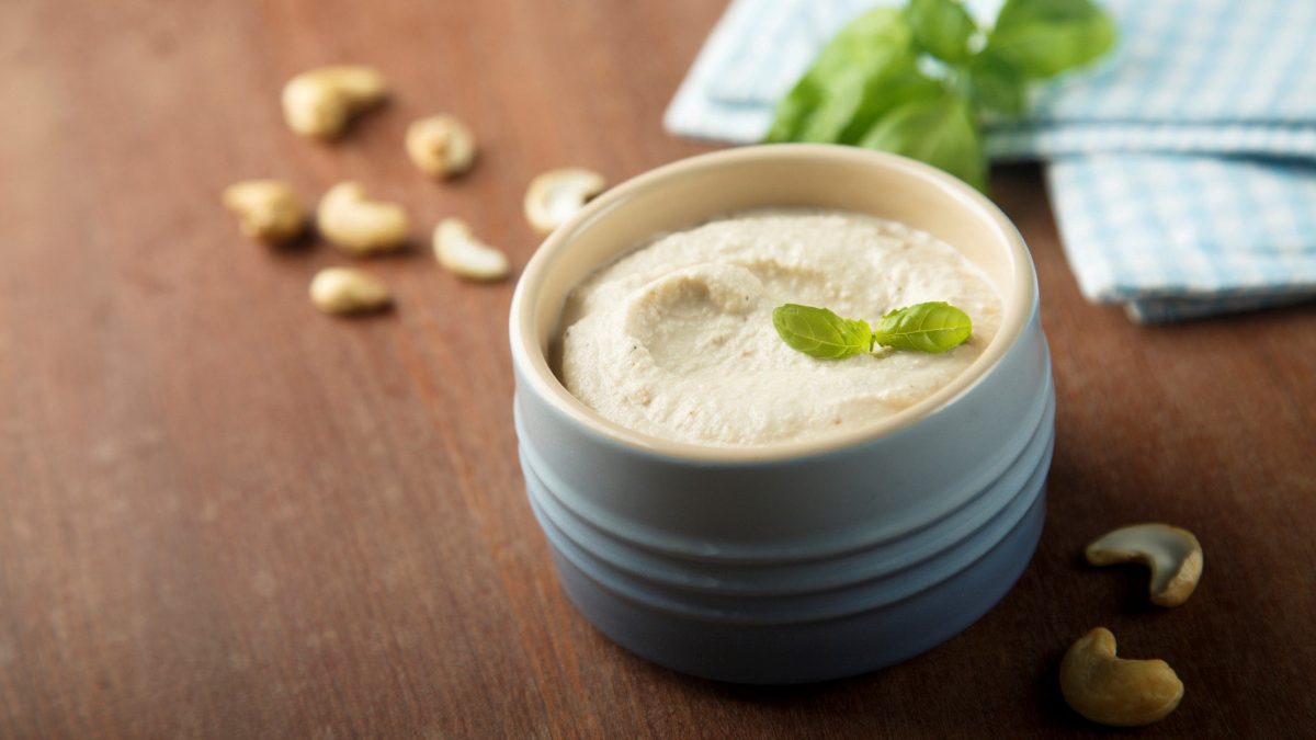 Cashew Creme in blauer Tonschale auf Holztisch. Daneben Cashews, Basilikum und blaues Tuch. Frontalansicht.