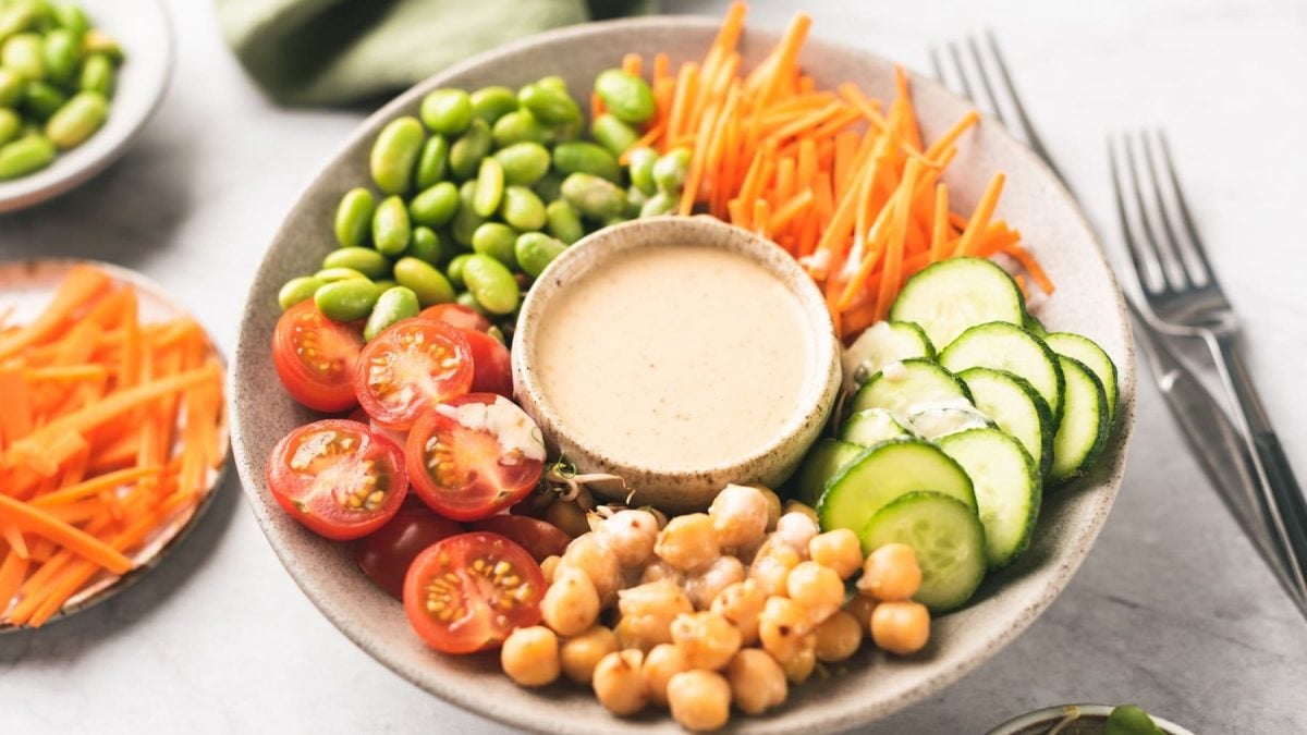 EIn Teller mit klein geschnittenen Karotten, Tomaten, Gurke, Kichererbsen und Edamame, in der Mitte steht ein Schälchen Cashew-Dressing.