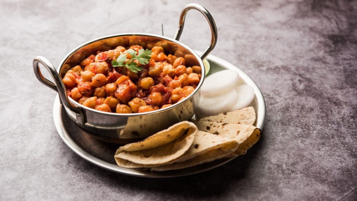 Chana Masala in silberner Schale auf Teller mit Brot auf grauem Untergrund. Draufsicht.
