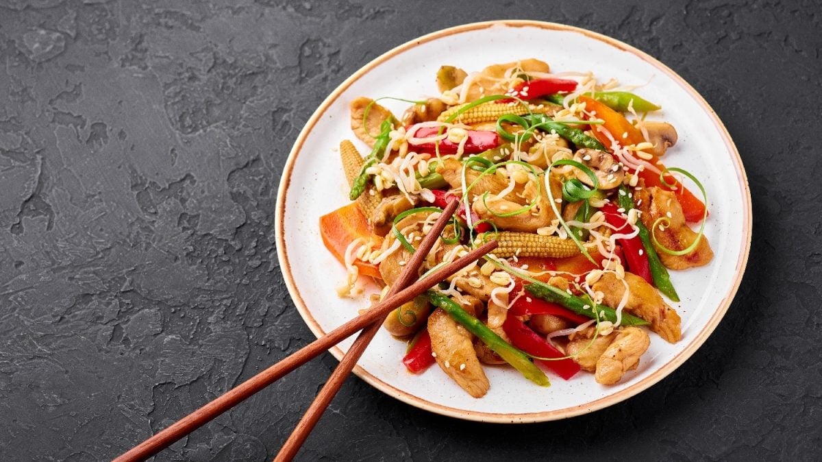 Ein weißer Teller mit dem Chop Suey auf einem schwarzen Untergrund und zwei Essstäbchen am unteren Tellerrand.