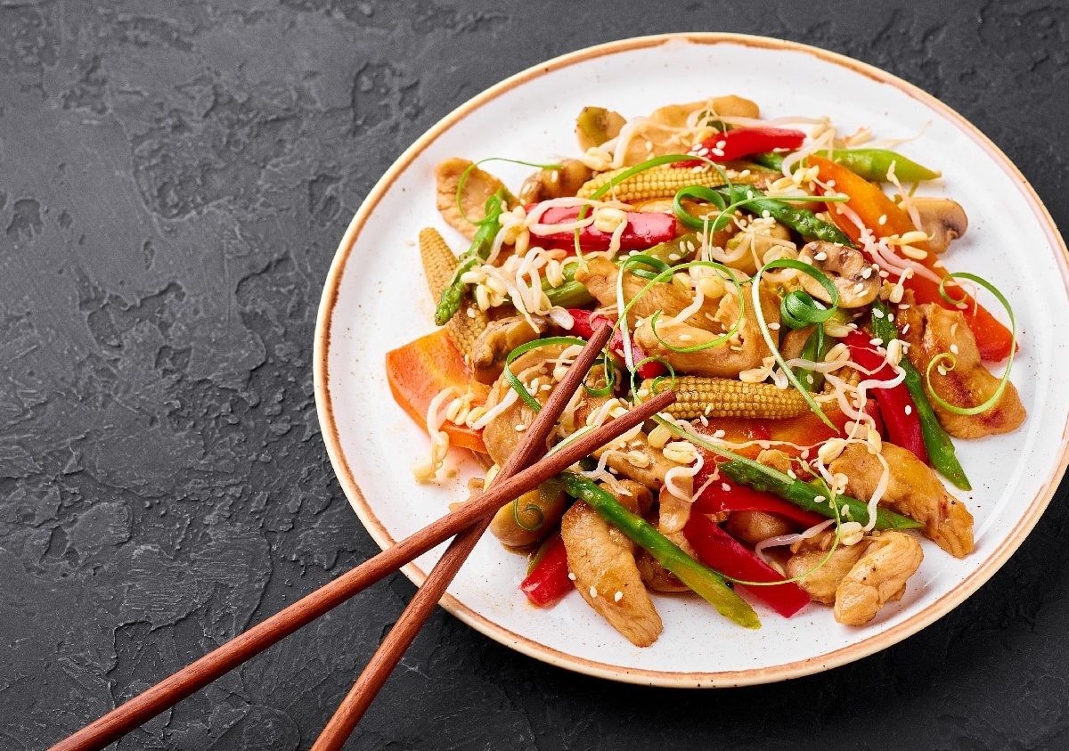 Ein weißer Teller mit dem Chop Suey auf einem schwarzen Untergrund und zwei Essstäbchen am unteren Tellerrand.