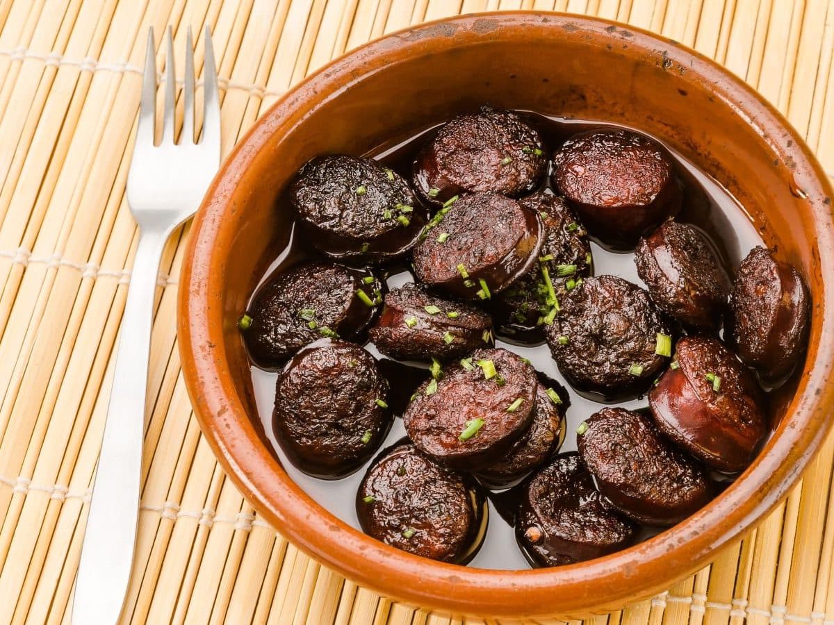 Chorizo in Rotwein in Tapasschale auf Bambusmatte. Daneben Gabel. Draufsicht.