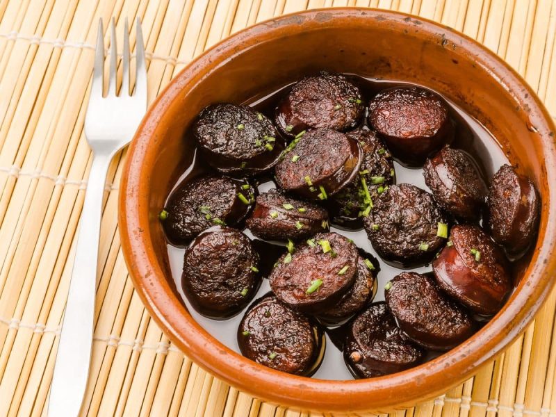 Chorizo in Rotwein in Tapasschale auf Bambusmatte. Daneben Gabel. Draufsicht.