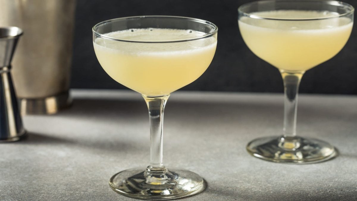 Zwei Corpse Reviver No. 2 auf grauem Tisch vor schwarzem Hintergrund. Dahinter Barmaß und Shaker. Frontalansicht