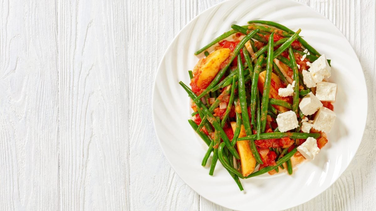 Ein weißer Teller mit grünen Bohnen, gekochten kartoffeln, Tomaten und feta steht auf einem weißen Untergrund. Das gericht heißt Fasolakia lathera.