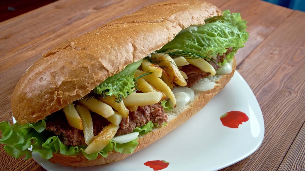 Streetfood Gatsby Sandwich mit Pommes und Masala-Steak auf weißem Teller und braunem Tisch. Frontalansicht.
