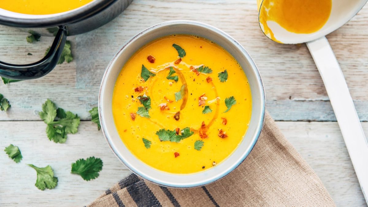Eine Schüssel gelbe Gemüsesuppe mit Petersilie und Chiliflocken garniert, daneben eine Kelle, Draufsicht.