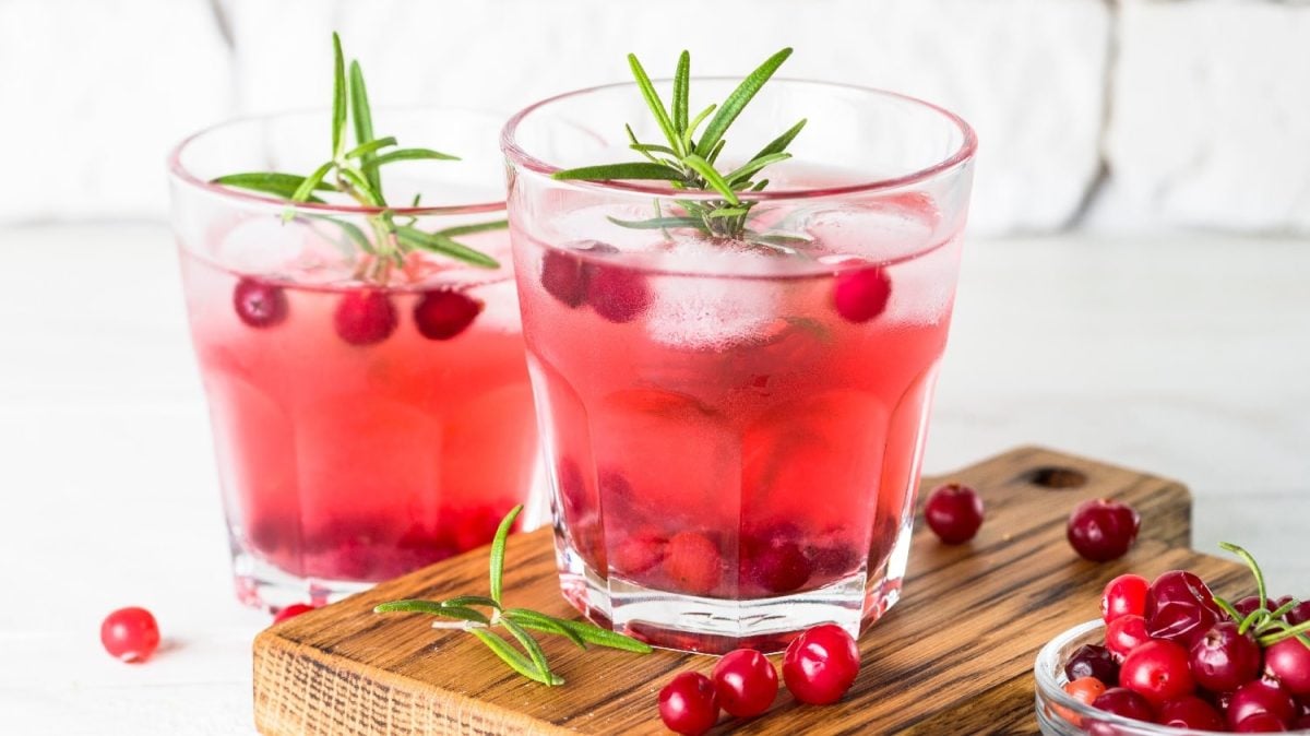 Zwei Gläser mit der Gin Fizz Kreation auf einem Holzbrett mit frischen Cranberrys und ein paar Rosmarinstielen vor einem weißen Steinhintergrund.