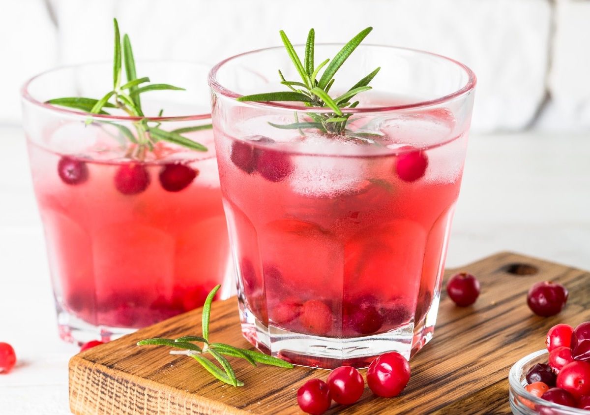 Zwei Gläser mit der Gin Fizz Kreation auf einem Holzbrett mit frischen Cranberrys und ein paar Rosmarinstielen vor einem weißen Steinhintergrund.