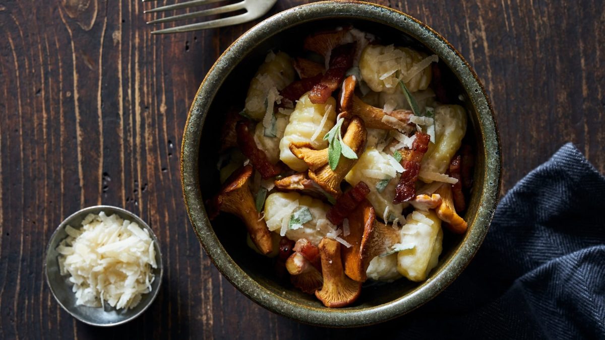 Eine graue Schüssel auf einem dunklen rustikalen Holztisch mit den Gnocchi und Pfifferlingen direkt von oben fotografiert. Daneben eine blaue Tischdecke und eine kleine Schale mit Parmesan auf der linken Seite.