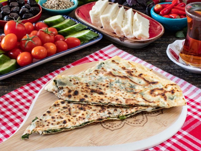 Gözleme auf Holzbrett auf rotem Tuch auf mit türkischen Speisen gedecktem Tisch. Daneben Tee. Draufsicht.