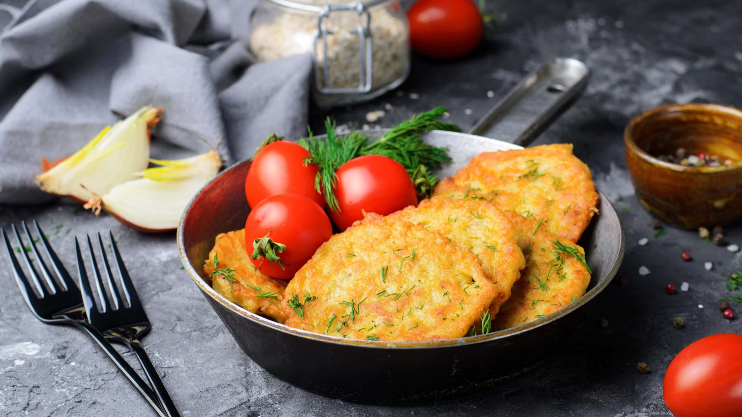 Ein dunkler tiefer Teller mit einigen Haferpuffern. Daneben ein paar Tomaten und eine angeschnittene Zwiebel auf einer dunklen Tischdecke.