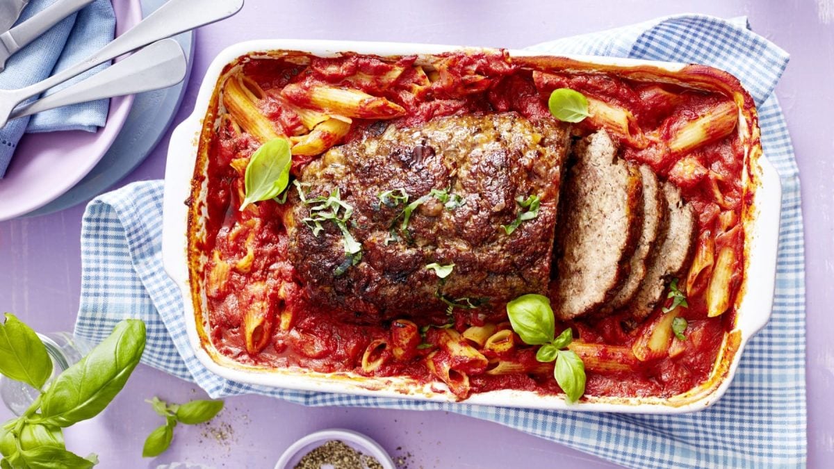 Italienischer One-Pot-Braten in weißer Auflaufform vor hellem Hintergrund, daneben Basilikumblätter und Geschirr