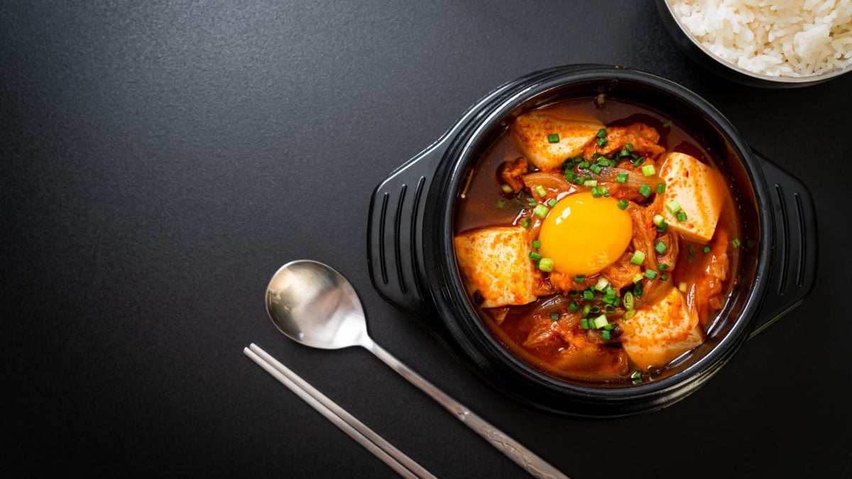 Ein schwarzer Topf mit Kimchi Jjigae neben Silberbesteck.