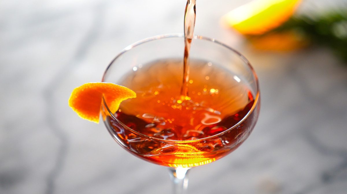 Knickerbocker Martini beim Eingießen in ein Martiniglas auf grauem Untergrund. Daneben Zitronenschale. Draufsicht.