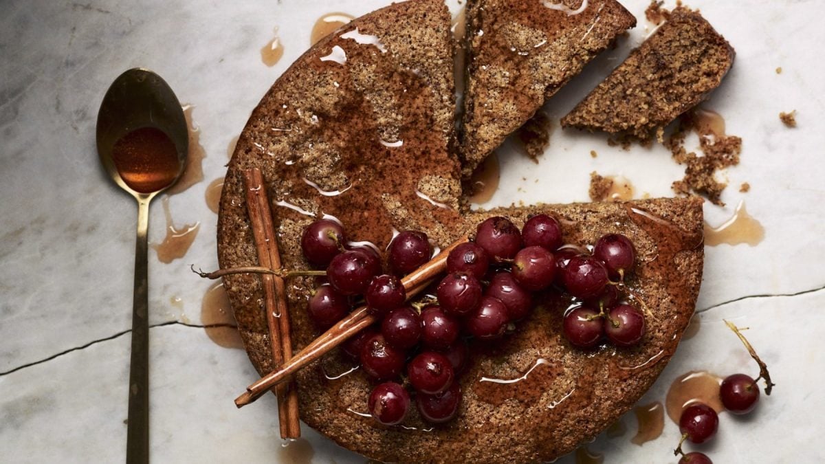 Runder Kokos-Haselnuss-Kuchen mit Trauben und Zimtstangen darauf neben einem Löffel mit Honig vor hellem Hintergrund