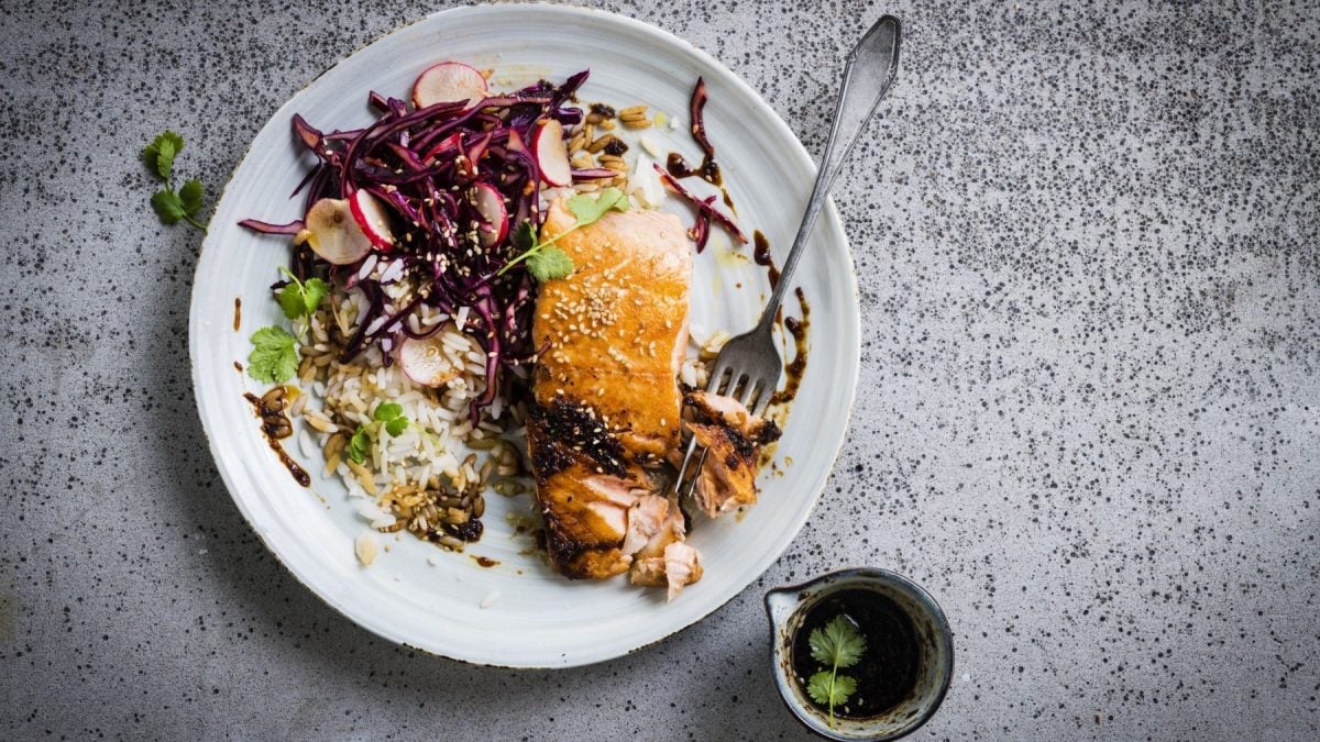 Lachs mit Rotkohl-Kimchi und Reis auf einem weißten Teller mit einer Gabel. Daneben steht ein Schälchen mit Sojasauce.