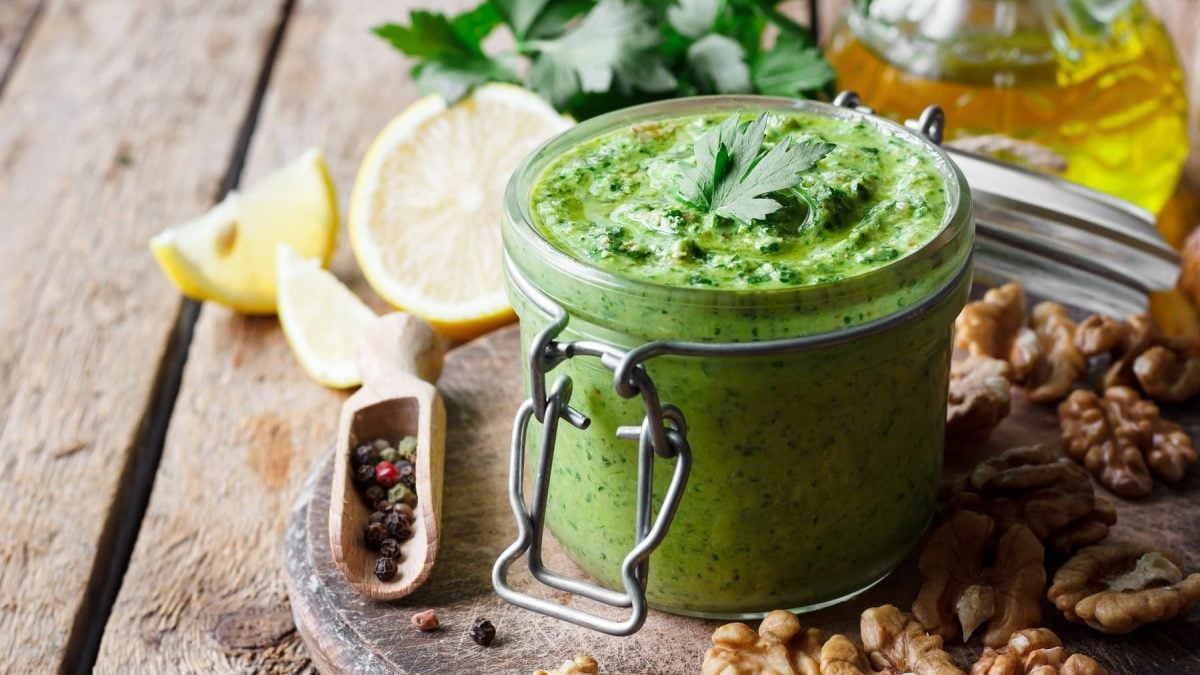 Ein Glas Mangold-Pesto, daneben liegen Zitronen, Walnüsse und bunter Pfeffer.