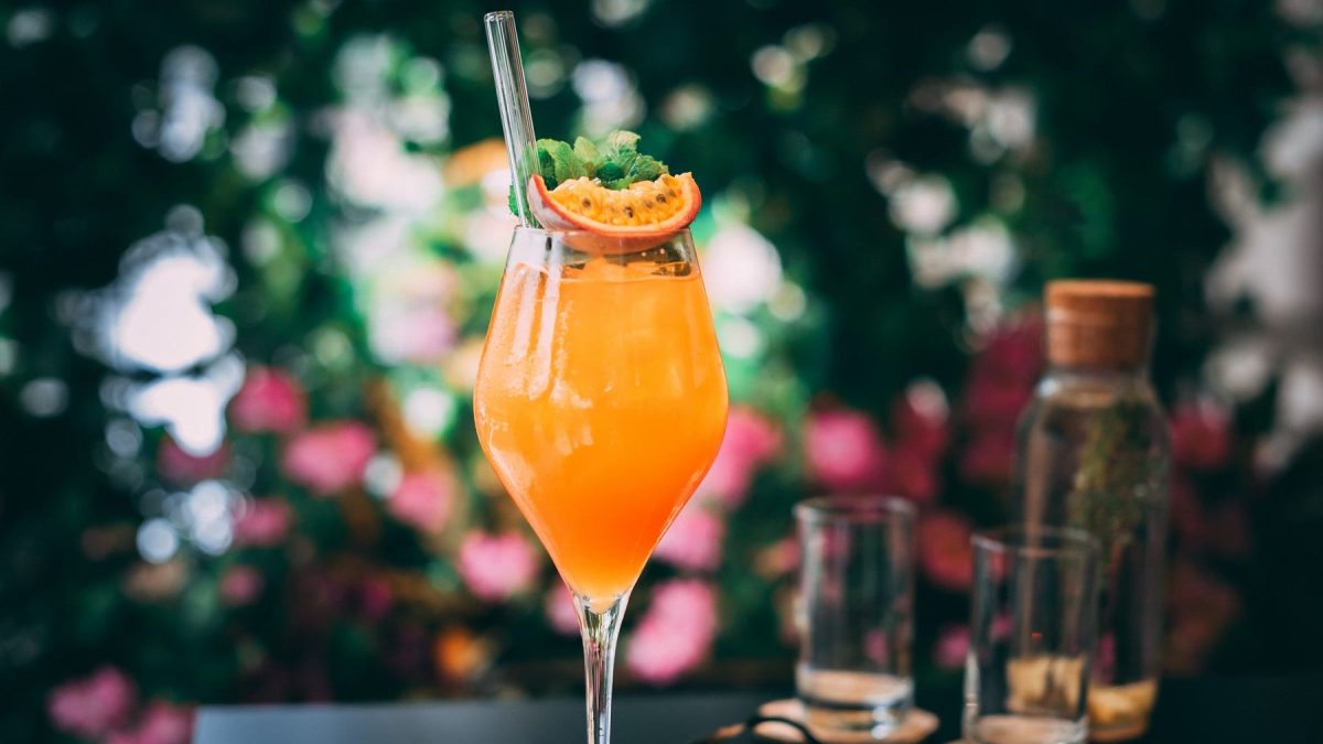Ein Weinglas mit Maracuja Spritz, garniert mit einer Maracuja-Scheibe und frischer Minze, darin ein Trinkhalm aus Glas.