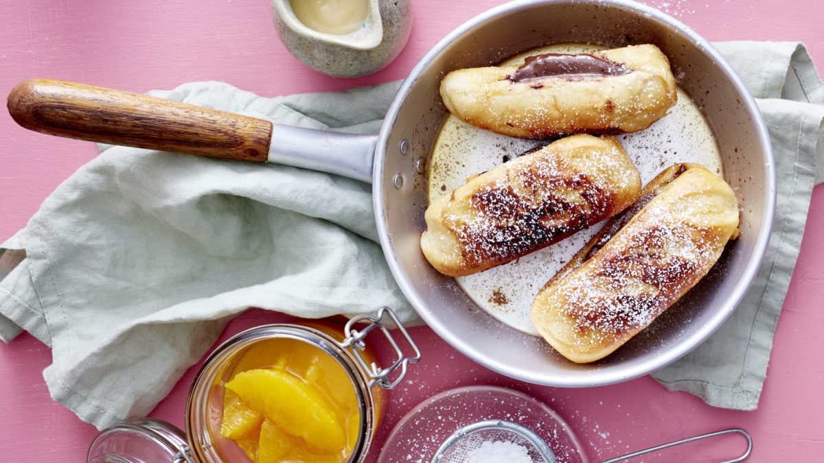 In einer Pfanne sind drei gefüllte Milchbrötchen mit Orangenkompott in einem Glas daneben vor rosa Hintergrund. Die Milchbrötchen sind gefüllt mit Nuss-Nougat-Creme und bestäubt mit Puderzucker.