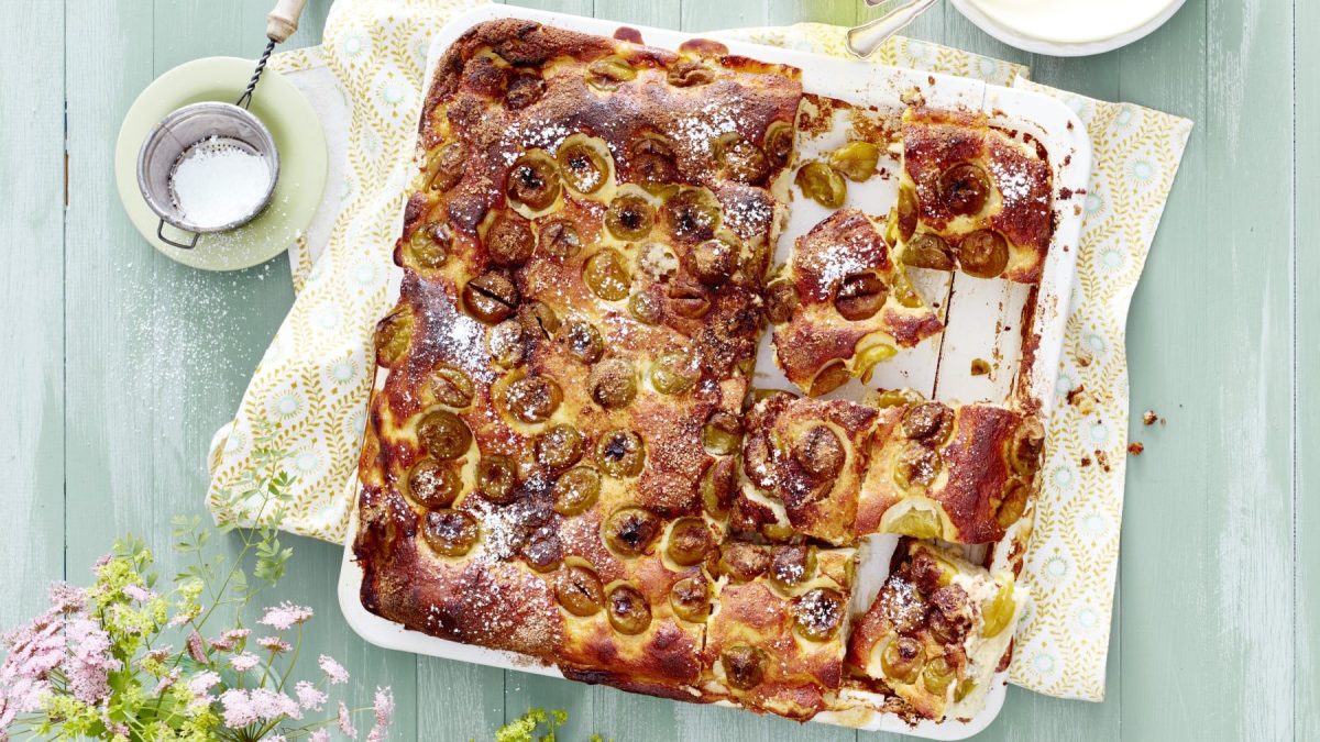 Mirabellen-Blechkuchen in weißer Ofenform auf einem Küchentuch, daneben Schüssel mit Sieb und Puderzucker vor hellem Hintergrund.