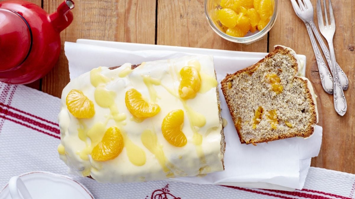 Mohn-Eierlikör-Kastenkuchen garniert mit Mandarinen auf Backpapier neben Geschirr und mehr Dosenmandarinen vor Holzhintergrund