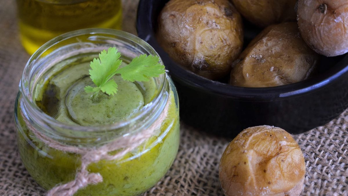 Mojo verde mit Paprika im Einmachglas auf hellem Untergrund. Daneben Patatas bravas und Olivenöl. Draufsicht.