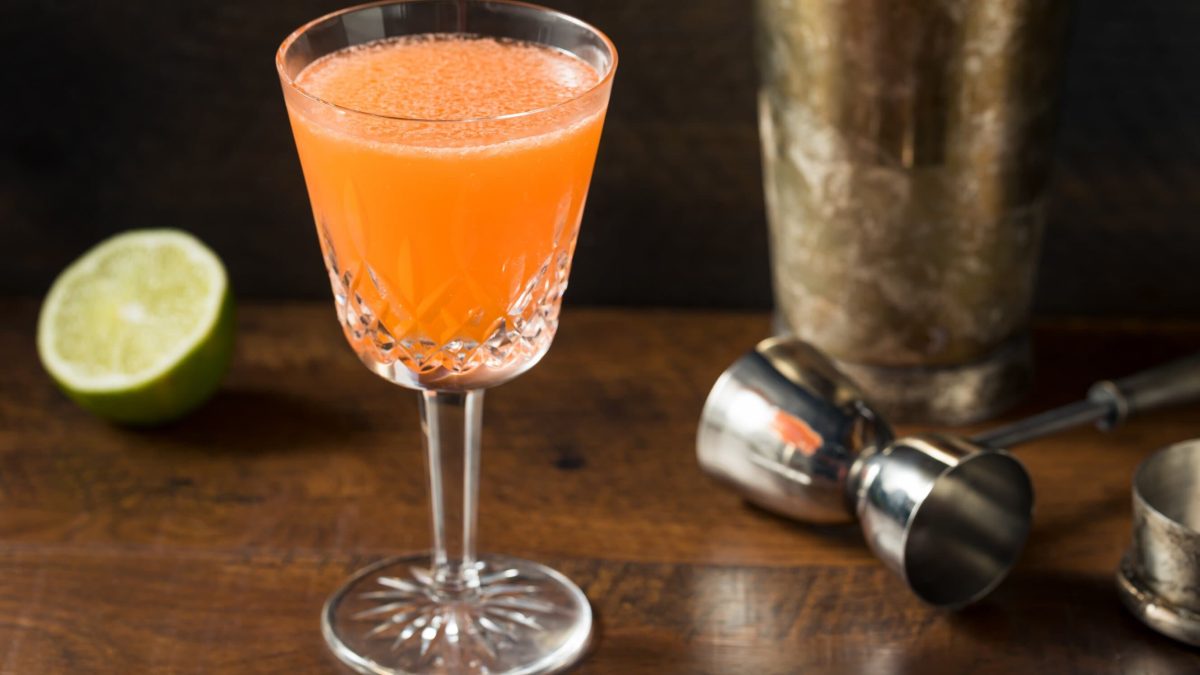 Naked and Famous Cocktail im Glas auf Holztisch. Im Hintergrund Shaker, Barmaß und Limette. Frontalansicht.