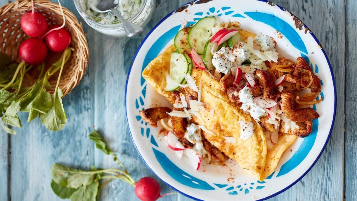Ein Omelett-Döner liegt auf einem weiß-blauen Teller. Daneben befinden sich verschiedene Zutaten.