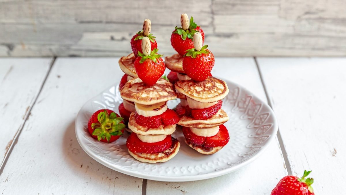 Pancake-Kebabs, auf Spieße aufgesteckte kleine Pancakes mit Erdbeer- und Bananenscheiben, auf einem Teller angeordnet.
