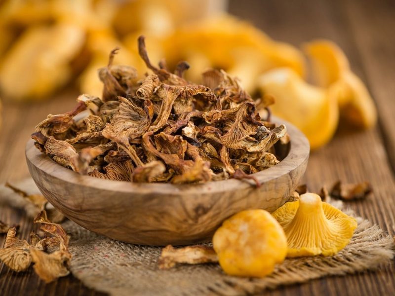 In einer Holzschale liegen getrocknete Pfifferlinge. Daneben liegen frische Pfifferlinge-
