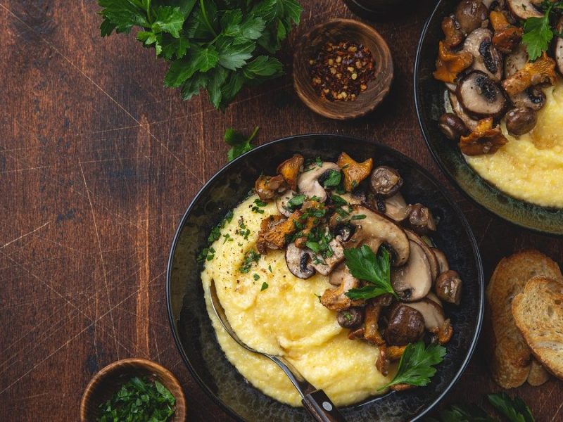 Zwei tiefe Teller Polenta mit gebratenen Pfifferlingen stehen auf einem Untergrund aus Holz. Daneben liegt Petersilie.