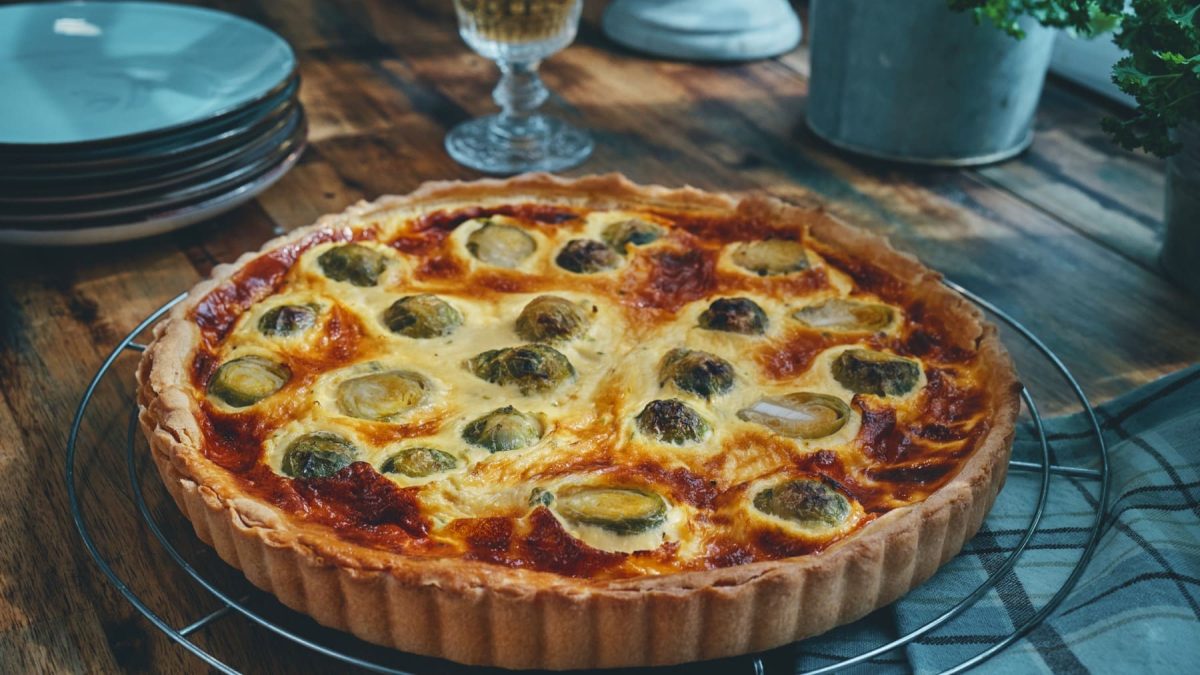 Eine Rosenkohl-Quiche liegt auf einem Gitter. Daneben steht Geschirr und ein Glas.