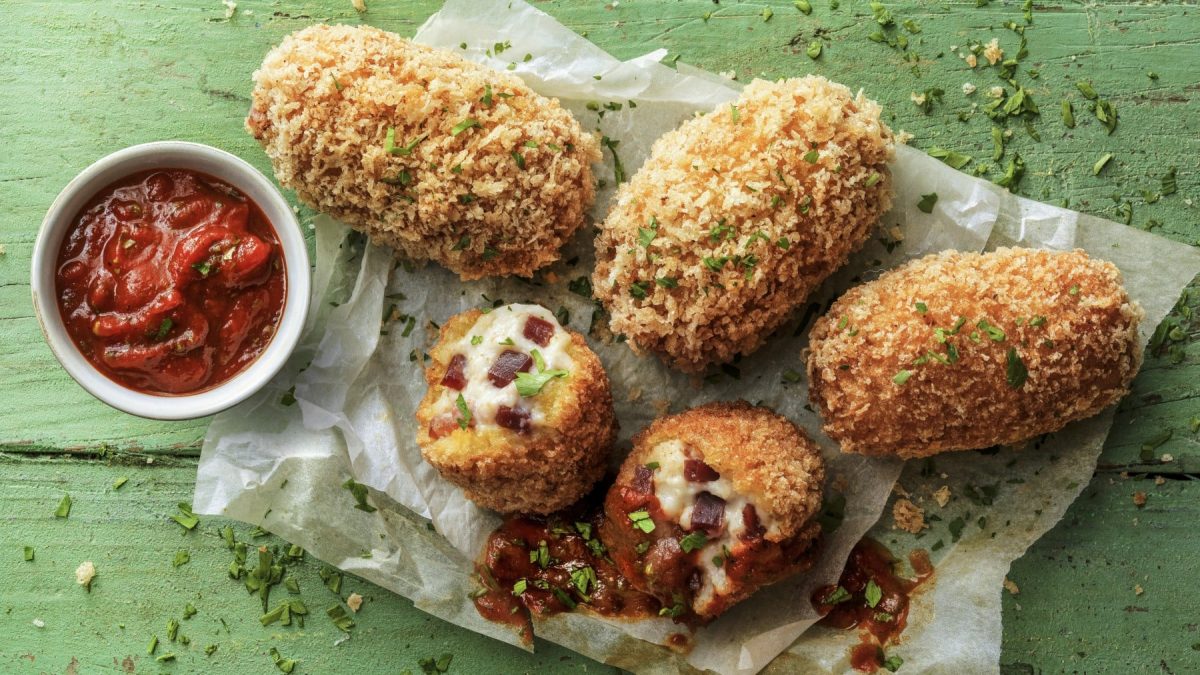 Fünf Schinkenkroketten auf Backpapier garniert mit Petersilie neben einer kleinen Schüssel Tomatensauce vor grünem Hintergrund