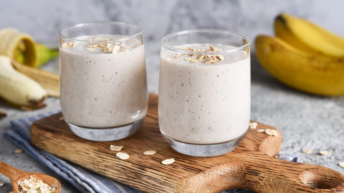 Zwei Gläser mit Bananen-Hafer-Smoothie auf einem Holzbrett neben frischen Bananen
