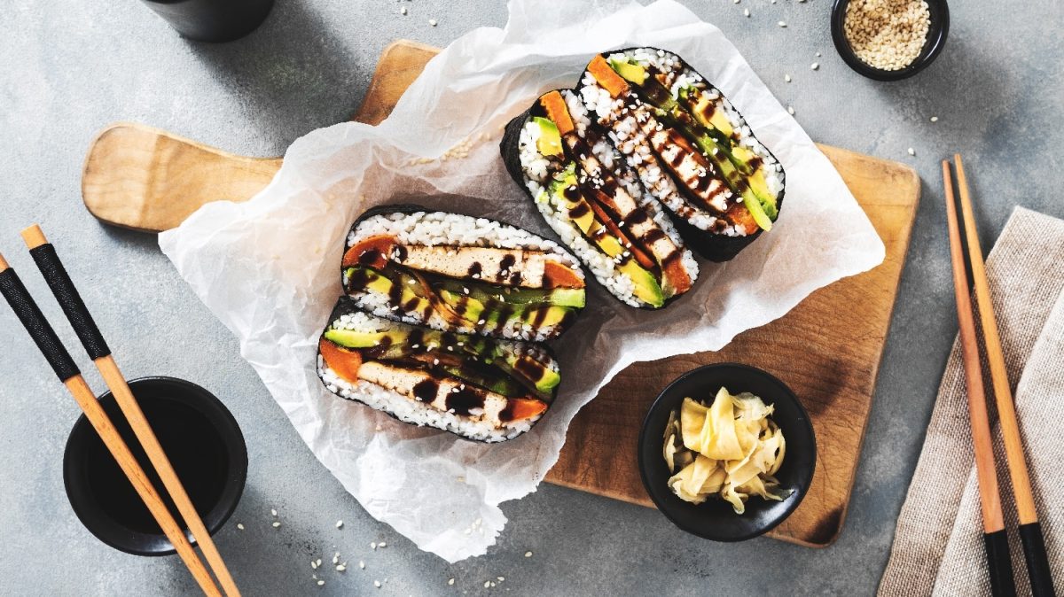 Ein aufgeschnittenes Sushi-Sandwich auf einem kleinen Holzbrett mit Essstäbchen, Sojasauce und Sesam.