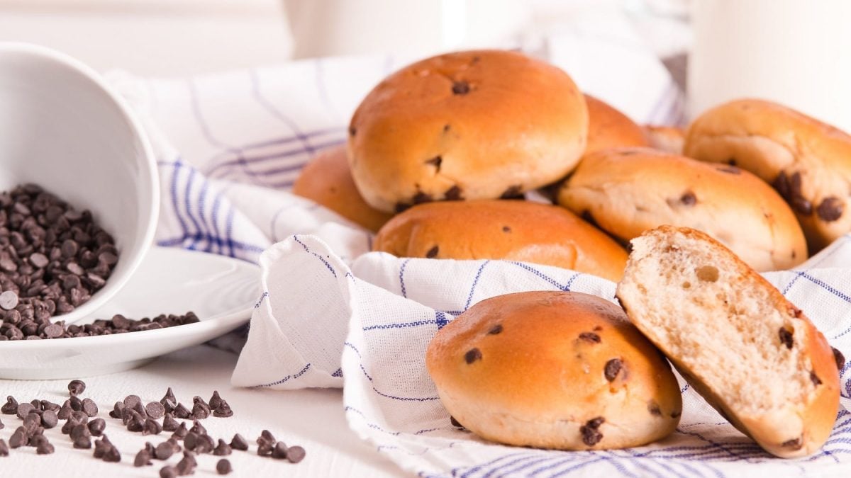 Vegane Schokobrötchen, eins davon angeschnitten, auf einem karierten Geschirrtuch, daneben eine umgekippte Schüssel mit Schokotropfen.