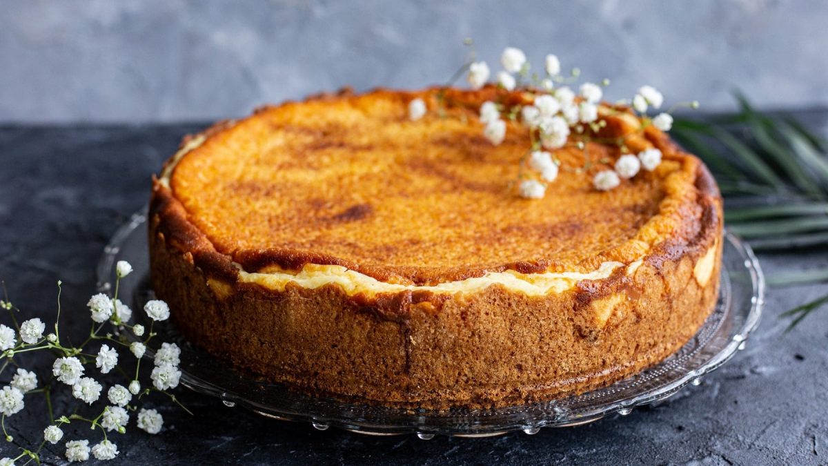 Ein veganer Mango-Vanille-Cheesecake auf einem Tortenteller aus Glas, mit weißen Blüten verziert.