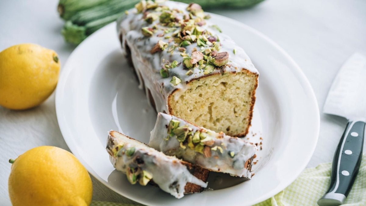 Ein angeschnittener Zitronen-Pistazien-Kuchen mit Puderzuckerguss und gehackten Pistazien, daneben liegen ein Messer und zwei Zitronen.