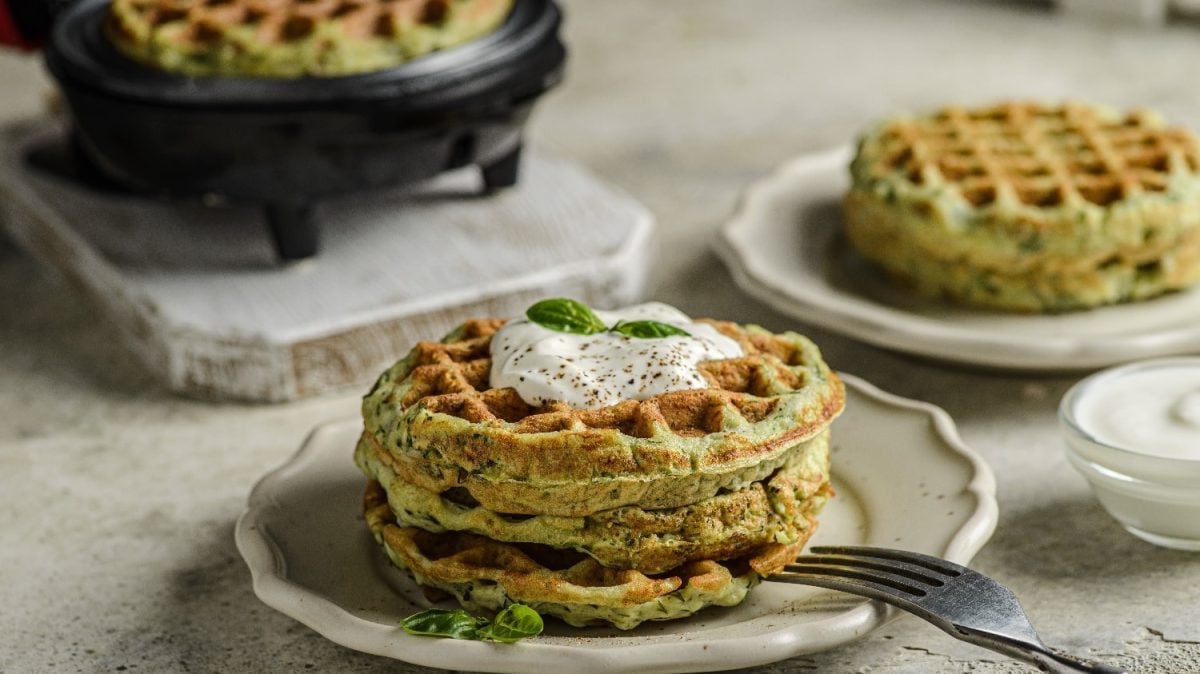 Zwei beige Teller mit jeweils zwei Zucchini-Waffeln auf einem beigen Tisch. Auf dem vorderen Teller ist außerdem ein Stück Burrata in der Mitter der Waffeln platziert.