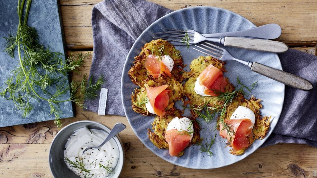 Zucchinipuffer mit Räucherlachs auf einem hellblauen Teller neben Dill und Schale mit saurer Sahne vor Holzhintergrund.